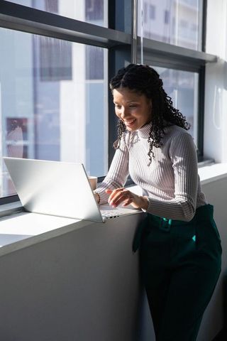 African american professional leaning on windowsill working on laptop in sunlit office