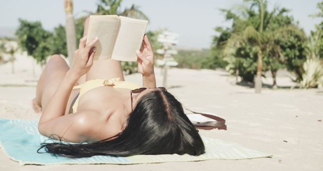 Hispanic woman in sunglasses lying on back on beach reading book, copy space, slow motion. Summer, relaxation and vacations.