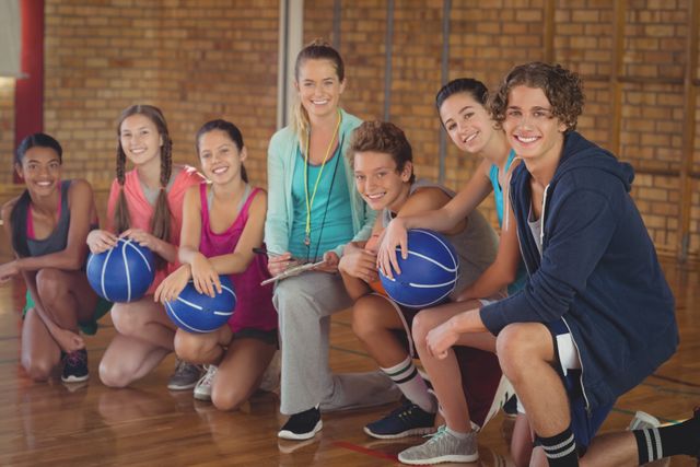 Female coach sitting with high school students in a gym, all holding basketballs and smiling. Ideal for use in educational materials, sports training guides, youth fitness programs, and promotional content for school sports events.