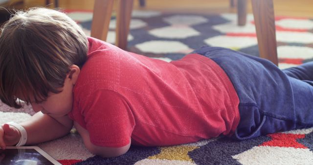 A young boy, dressed in a red shirt and blue pants, lying on a colorful carpet while using a tablet device at home. This image can be used to illustrate topics related to children's technology use, digital education, remote learning, or home entertainment. Ideal for use in articles, educational materials, and advertisements targeting parents and educators.