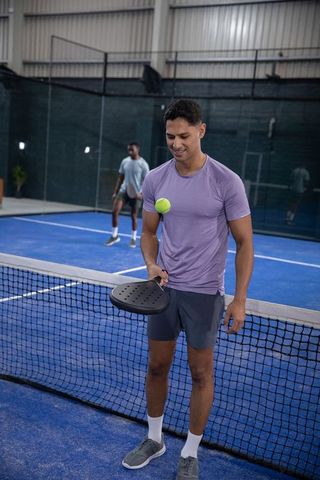 Two men playing padel at net on indoor blue court balancing ball on paddle, training together
