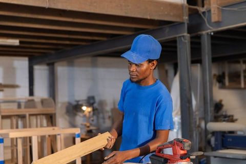 African american carpenter inspecting wooden plank at workshop workbench with sander