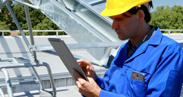 Engineer in blue uniform examines a tablet at a construction site. His focus on technology suggests modern approaches in the industry.