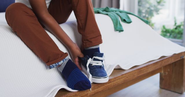 This image shows a close-up of a person sitting on a wooden bed, putting on a blue sneaker. Ideal for themes related to daily routines, casual fashion, home interior, and relaxed lifestyles. Can be used in blogs, advertising campaigns for casual footwear, and lifestyle magazines.