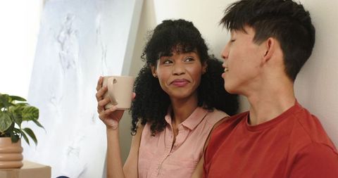Cozy multicultural couple chatting at home while holding coffee mug and smiling warmly