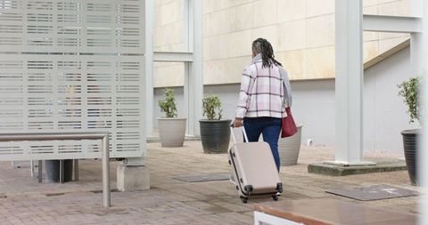 African American Woman Walking With Rolling Suitcase Carrying Red Tote On Urban Plaza