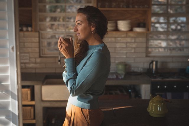 Caucasian woman standing by window, holding coffee cup, enjoying morning light. Ideal for lifestyle blogs, relaxation themes, home decor inspiration, morning routines, and wellness content.