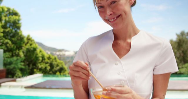 Smiling woman in white uniform stirring an orange smoothie by the poolside. Background features lush greenery and swimming pool, suggesting a relaxing and refreshing summer setting. Perfect for use in wellness, spa, summer vacation, healthy living, and outdoor leisure contexts.