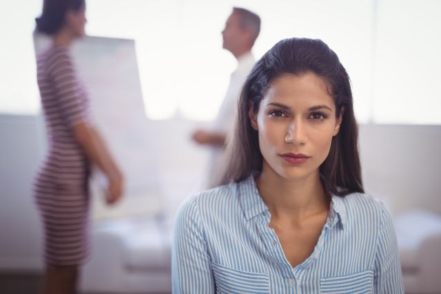 Confident businesswoman standing in foreground with colleagues discussing in background. Ideal for use in articles about leadership, workplace dynamics, professional development, and corporate environments.