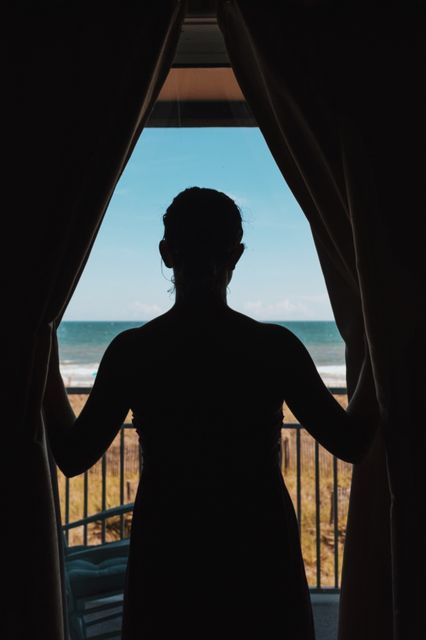 Silhouette of woman standing by window looking out at ocean scene, suggesting contemplation and relaxation. Could be used for advertisements related to vacation rentals, travel, mindfulness and wellness, or illustrating concepts of relaxation and tranquility.