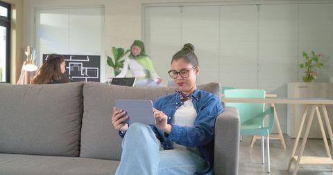 Young professional using tablet on sofa while team collaborates in modern coworking lounge