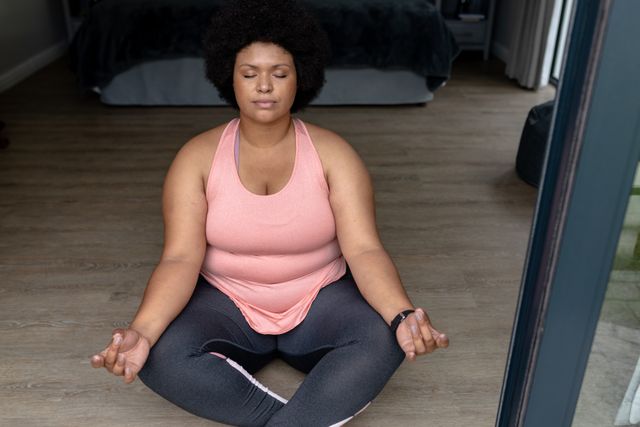 African American mid adult woman sitting in a meditation posture with eyes closed at home entrance. Ideal for use in content related to wellness, mindfulness, fitness, active lifestyle, mental health, and peaceful living. Perfect for promoting yoga and relaxation practices.