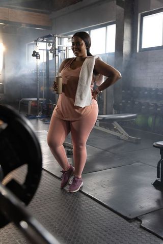 Confident african american woman working out in gym weight area holding shaker and towel