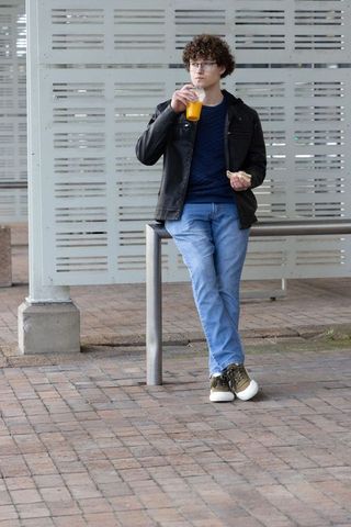 Young man leaning on rail at urban shelter sipping orange juice and eating pastry in casual outfit