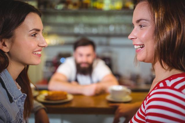 Two female friends are sitting at a cafe counter, smiling and interacting with each other. A barista is seen in the background. This image can be used for promoting social gatherings, coffee shops, friendship, and leisure activities.