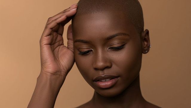 Holding hand to temple, closing eyes, shaved-head closeup with gold eyeshadow and stud earring