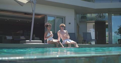 Diverse couple relaxing by modern villa pool, sipping drinks with feet in turquoise water