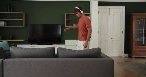 Young man with headphones standing by sofa watching TV in modern minimalist living room