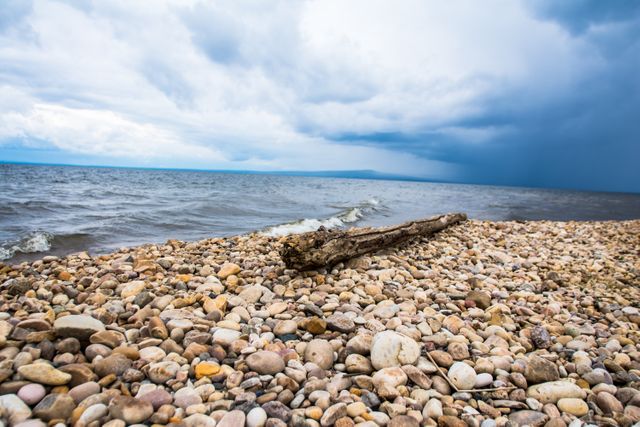 Beach with multicolored pebbles, sea and horizon. Driftwood in foreground, stormy clouds in sky. Suitable for nature scenes, weather themes, coastal and seaside photography.