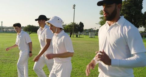 Four cricket players walking across sunlit park field wearing white uniforms and caps