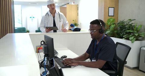 Clinic receptionist typing at desk with headset and doctor consulting tablet in background