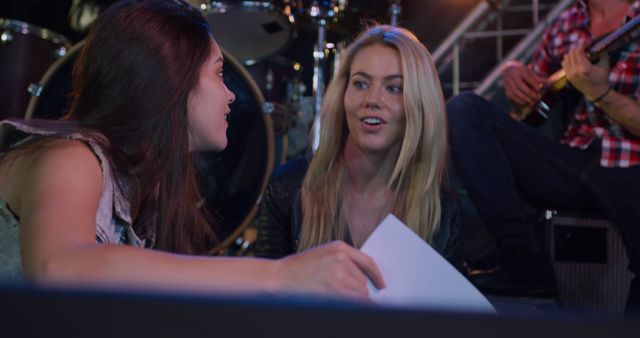 Two women discussing and planning during a band rehearsal. Drums and guitars visible in the background, indicating a musical setting. The women seem engaged and serious about their collaboration, with one holding a piece of paper. This could be used in articles about teamwork, women in music, or collaboration in creative settings, and marketing materials for music-related events or workshops.