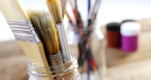 Paintbrushes are arranged in a glass jar on a wooden surface, with blurred paint containers in the background, with copy space. Art supplies like these are essential for artists and hobbyists to create various paintings and crafts.