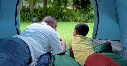 African american father and son bonding in backyard tent camping together sharing smartphone