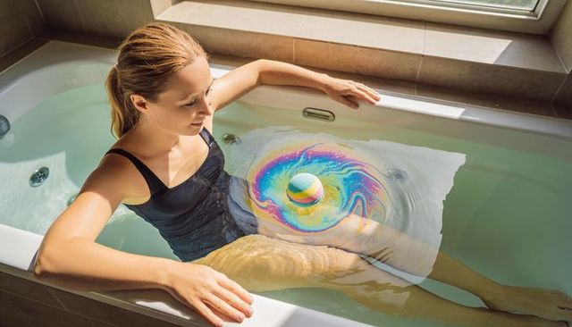 Woman soaking in bathtub with rainbow bath bomb creating colorful swirl