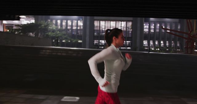 Side view of a fit Caucasian woman with long dark hair wearing sportswear, exercising outdoors in the city during the night, running through the street.