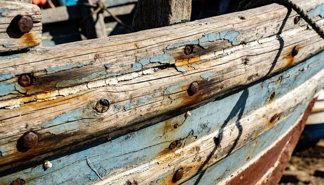 Weathered wooden boat hull revealing peeling blue paint, rusted bolts, rope shadow texture
