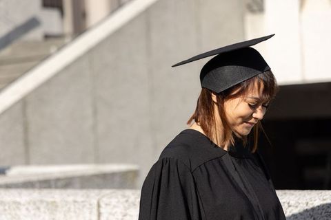 Asian graduate woman wearing mortarboard and black gown standing by concrete campus ledge