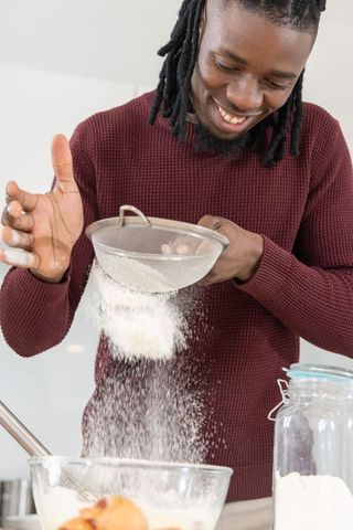 African american man sifting flour in modern kitchen smiling while baking homemade treats