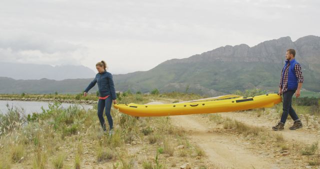 Man and woman carry bright yellow kayak along rough trail near serene mountain lake. Ideal for outdoor adventure, teamwork, hiking promotions, nature excursion concepts, travel blogs, and recreational activity websites.