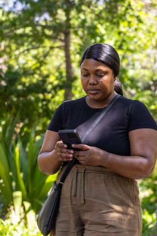 African woman using smartphone in park wearing black t-shirt and tan pants