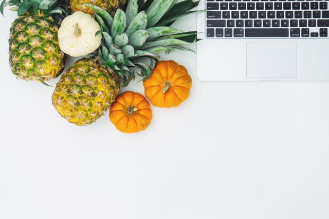 The photo shows pineapples and small pumpkins next to a modern laptop on a clean white surface. This image is suitable for promoting healthy lifestyles, modern workspaces, minimalistic decor, or autumn-themed events. It can be used in blogs, social media, advertisements for health products, or office settings.