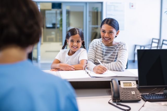Mother and daughter are at hospital reception desk, filling out paperwork. They are smiling, indicating a positive and welcoming environment. This image can be used for healthcare, family care, medical services, and hospital-related content.
