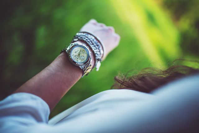 Close-up focusing on luxury watch worn on woman's wrist, showing elegant silver design with a blurred green outdoor background. Great for marketing high-end fashion accessories, timepieces, jewelry, or lifestyle blogs. Also suitable for illustrating themes of time management, style, and elegance.