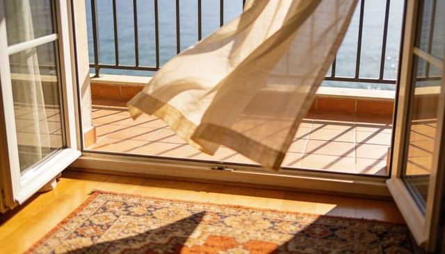 Swaying sheer curtain over open balcony doors with sunlit terracotta tiles and sea view