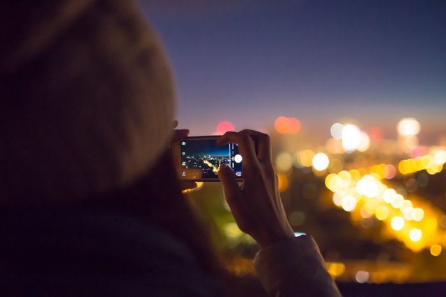 Person in beanie capturing vibrant city skyline at night with smartphone. Enhances technology's role in everyday life, suitable for travel, urban life themes, and technology-related content depicting capturing moments and cityscapes at night.