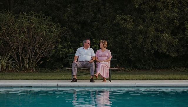 Mature couple relaxing on bench by backyard pool, sharing evening conversation