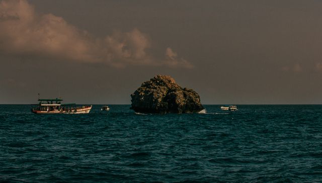 Boats are navigating in the calm waters around a small rocky island during sunset, creating a peaceful and serene scene. The sky is tinged with warm colors, adding to the tranquil atmosphere. This image is suitable for travel, adventure, and nature-themed content, showcasing the beauty of marine landscapes and boating experiences at twilight.