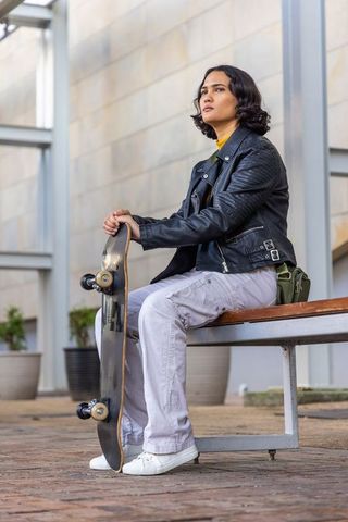 Sitting with skateboard and green shoulder bag wearing leather jacket in urban plaza