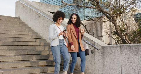 Diverse young women walking campus stairs, chatting, using smartphone, holding coffee