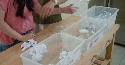 Roommates sorting recyclables into labeled paper, glass and plastic bins in kitchen