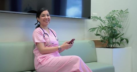 Smiling nurse in pink scrubs sitting on clinic bench holding smartphone and wearing stethoscope