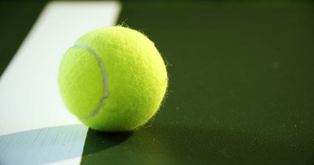 This image showcases a close-up view of a tennis ball lying on the baseline of a green tennis court, captured under natural lighting. Perfect for use in sport-related promotions, tennis coaching websites, training manuals, or illustrating articles and blogs about tennis matches and competitions.