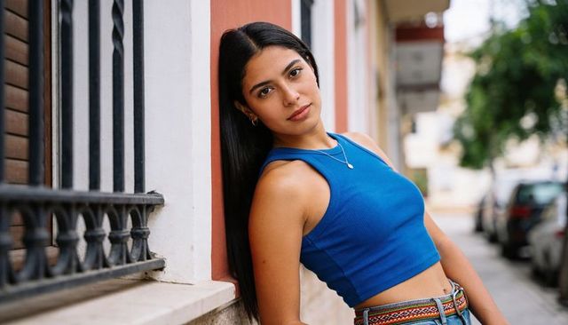 Leaning woman on urban facade wearing blue crop top, high-waist jeans and colorful belt