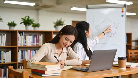 Student writing notes while tutor pointing at network diagram on whiteboard in library