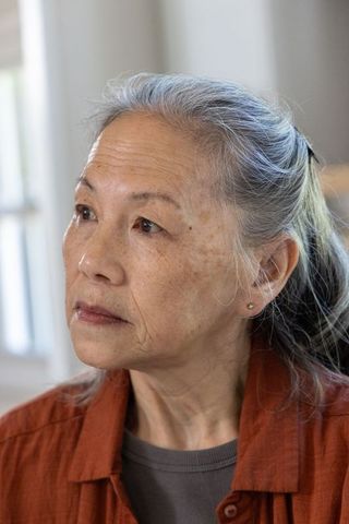 Senior asian woman gazing thoughtfully upward left wearing rust shirt, interior portrait